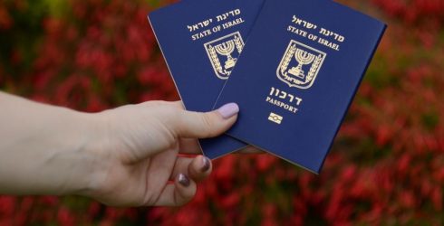תמונת נושא של המאמר על עורך דין הגירה לישראל בה מוצגת יד של אישה מחזיקה שני דרכונים ישראליים כחולים פתוחים למחצה על רקע שיח אדום