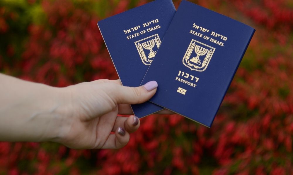 תמונת נושא של המאמר על עורך דין הגירה לישראל בה מוצגת יד של אישה מחזיקה שני דרכונים ישראליים כחולים פתוחים למחצה על רקע שיח אדום