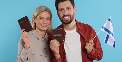 תמונת נושא של המאמר ויזת עבודה בישראל מדריך מלא המציגה זוג מחייך אישה בלונדינית וגבר מזוקן מחזיקים דרכונים חומים ודגל ישראל על רקע כחול בהיר המייצגים עליה או הגירה